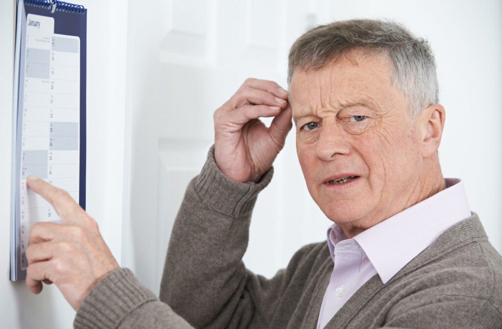 A senior man with a look of confusion scratches his head as he tries to remember something while pointing to a specific date on the wall calendar.