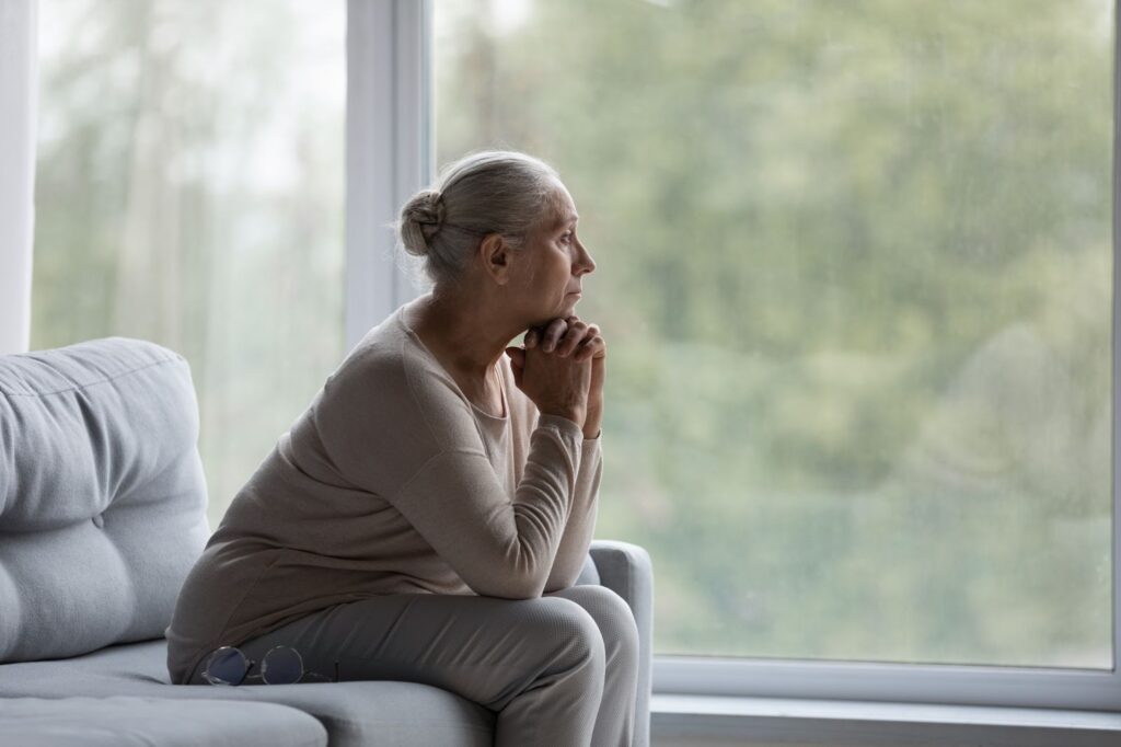 A pensive older woman with dementia sits at home and stares out the window