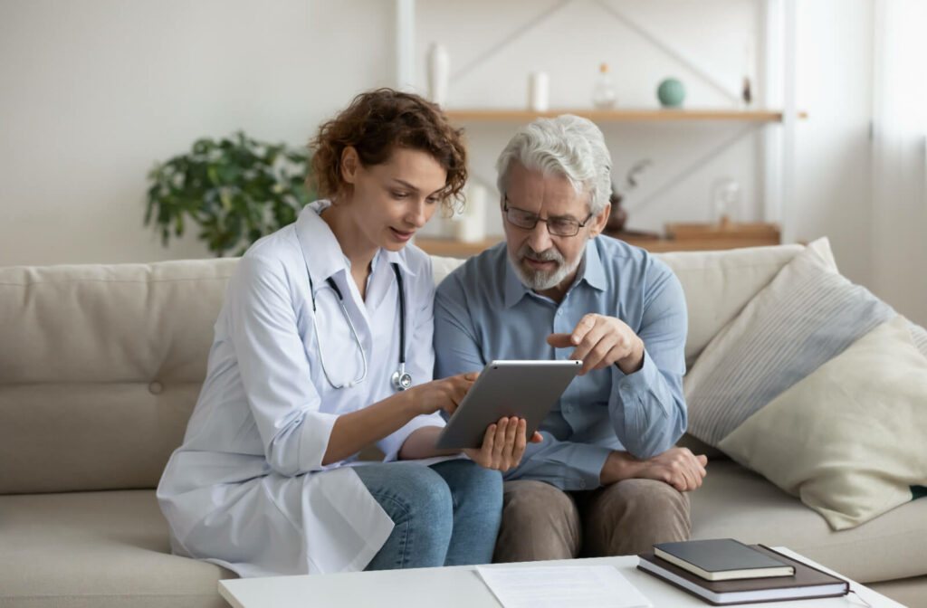 A caregiver in memory care sitting on the couch beside an older adult with Parkinson's, outlining their new care plan on a tablet.