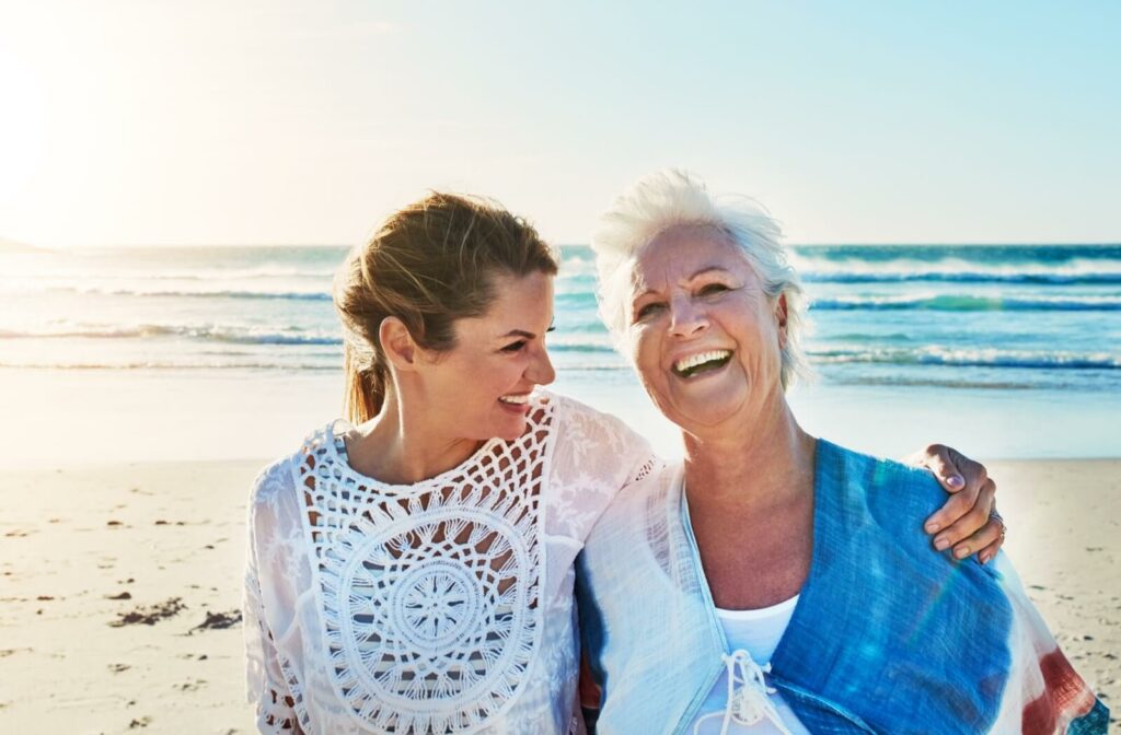 An adult and their senior parent smiling on a white sand beach with rolling waves in the background