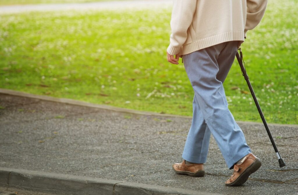 A senior with a walking cane walks along a cement sidewalk and green grass outdoors on a nice day