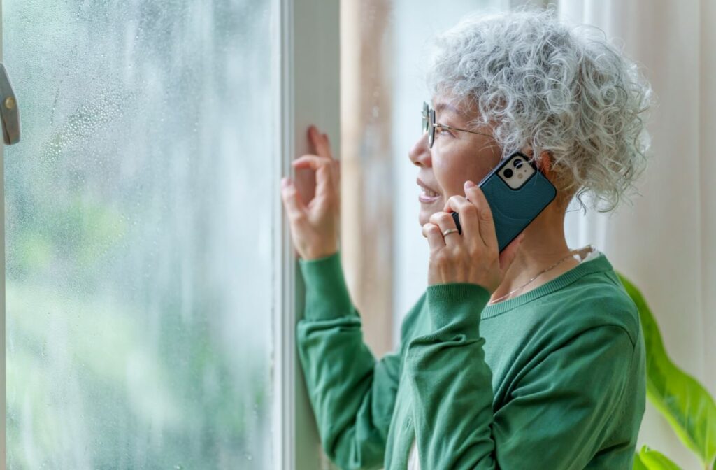 A senior, on the phone with a loved one, worriedly looks outside at the pouring rain from a hurricane passing through